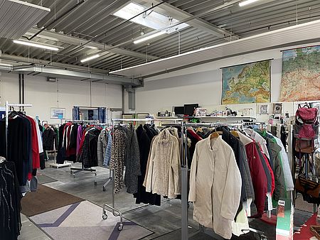 Man sieht Stangen mit Kleidung, insbesondere Winterjacken, im Göttinger Spendenzentrum des DRK. Im Hintergrund ist eine Weltkarte an der Wand und diverse Gegenstände erkennbar, die dort gespendet wurden und verkauft werden