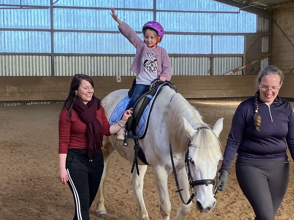 Reitstallausflug der DRK-Kita Scheden: Ein Kind reitet auf einem weißen Pferd in der Reithalle, daneben laufen zwei Erwachsene Personen