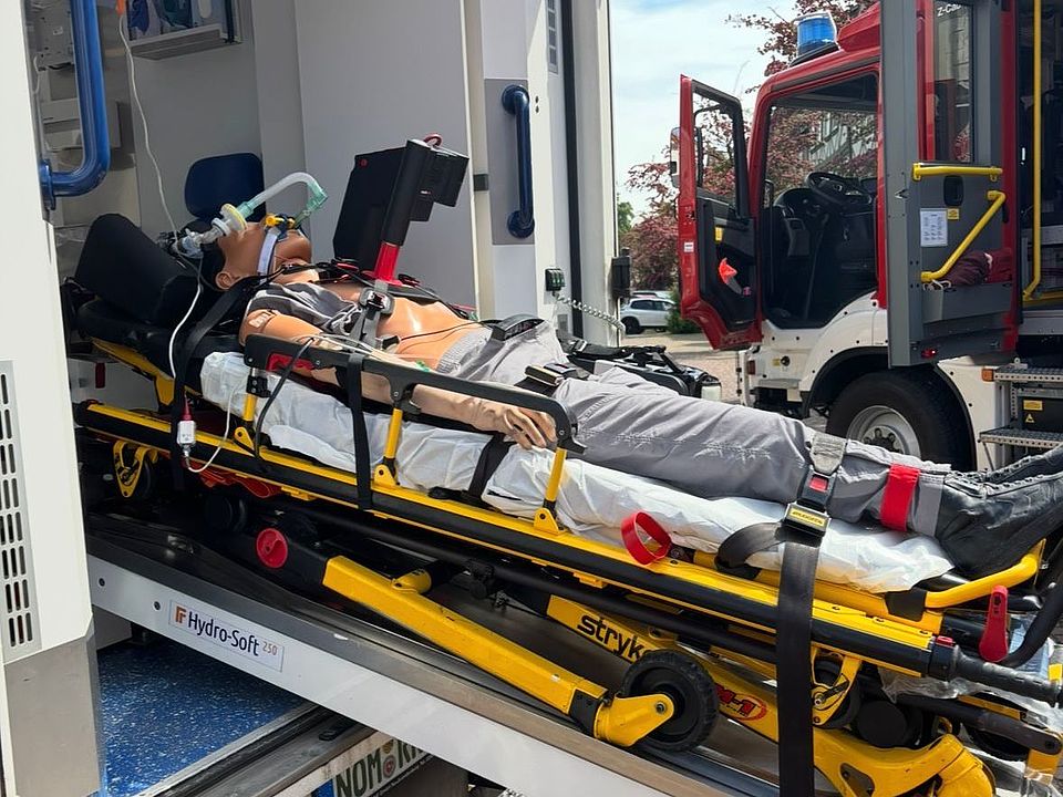 Eine Trage mit Übungspuppe wird halb aus einem Rettungswagen herausgefahren und veranschaulicht die Abläufe im Einsatz. Im Hintergrund steht ein Feuerwehrfahrzeug, während die Szene Teil einer Demonstration des DRK-Rettungsdienstes auf dem Bienenmarkt Uslar ist.