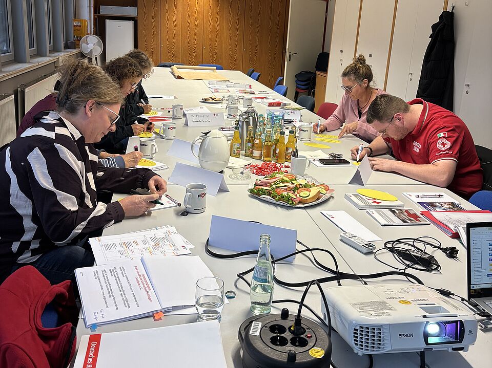 2026_10_23_DRK_Workshop_in_Northeim__4_.jpeg Die Teilnehmenden sitzen an einem Konferenztisch und bearbeiten Arbeitsaufträge. Auf dem Tisch liegen Schulungsunterlagen, Moderationsmaterialien und Verpflegung für den gemeinsamen Austausch.