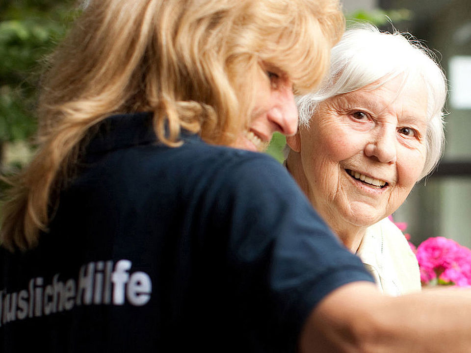 2025_12_10_Hauswirtschaftliche Hilfe_FUD_offiziell_Zelck Eine ältere Frau mit weißem Haar schaut freudig in die Kamera und im Vordergrund steht eine jüngere Frau, die die ältere Frau unterstützt. Man sieht sie nur von hinten seitlich