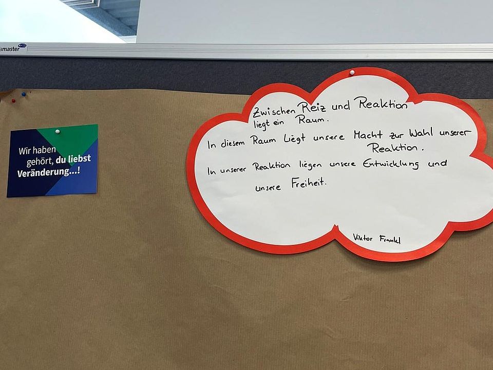 Im Konferenzraum des DRK‑Kreisverbandes Göttingen‑Northeim e. V. hängt an einer Pinnwand ein großes braunes Papier, das für eine Gruppenarbeit vorbereitet wurde. Rechts oben auf dem Papier ist eine große Wolkenform aus Pappe befestigt, weiß mit rotem Rand. Darauf steht handgeschrieben ein Zitat von Viktor Frankl: „Zwischen Reiz und Reaktion liegt ein Raum. In diesem Raum liegt unsere Macht zur Wahl unserer Reaktion. In unserer Reaktion liegen unsere Entwicklung und unsere Freiheit.“ Links daneben ist eine kleine farbige Postkarte mit dem Text „Wir haben gehört, du liebst Veränderung…!“. Am oberen Rand des Bildes ist ein Fensterrahmen des Konferenzraums sichtbar.