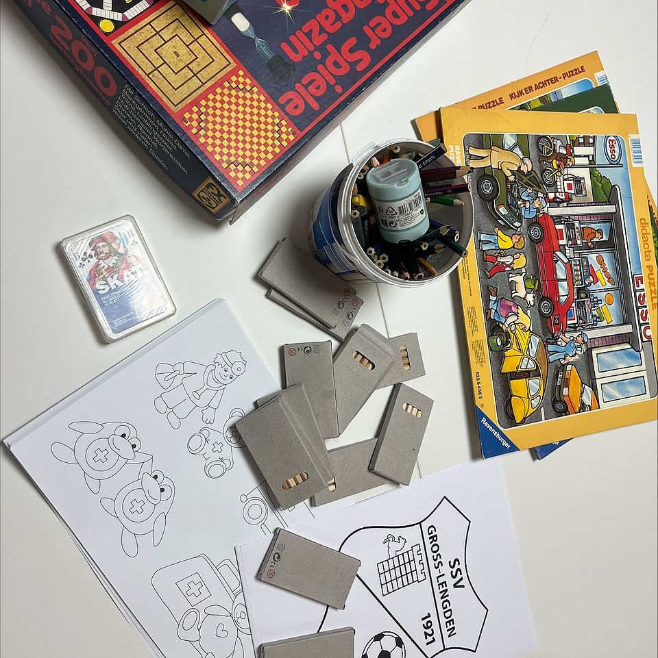 2026_02_Blutspende_Groß_Lengden__3_.jpeg Auf einem Tisch liegen verschiedene Spiel‑ und Bastelmaterialien ausgebreitet. Links befinden sich mehrere bemalte Ausmalvorlagen mit Tiermotiven sowie ein Papier mit einem Vereinswappen. Daneben liegen mehrere rechteckige graue Kartonstücke. In der Mitte steht ein Becher mit Stiften und kleinen Bastelutensilien. Rechts davon liegen mehrere bunte Kinderpuzzles mit Fahrzeug‑ und Werkstattmotiven. Oben im Bild sind zwei große Spielesammlungen in Schachteln zu sehen. Die Szene zeigt eine zusammengestellte Auswahl an Beschäftigungsmaterialien, wie sie bei einer Kinderbetreuung oder Veranstaltung genutzt werden, hier bei der Blutspende des DRK.
